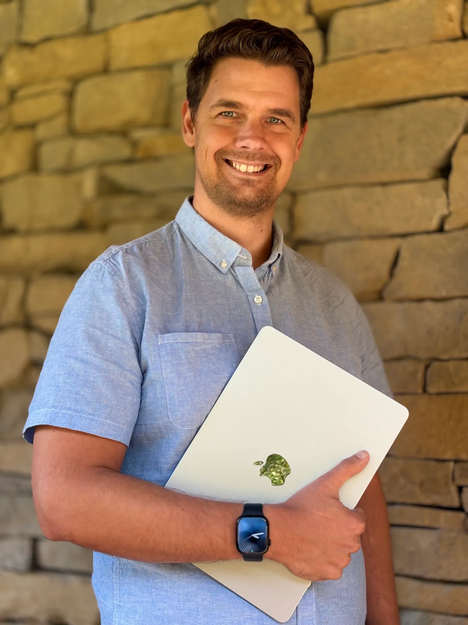 Filip ve světle modré košili se usmívá a drží notebook před kamennou zdí